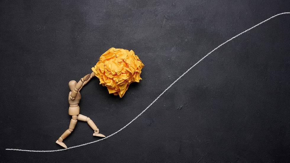 Wooden mannequin pushes large crumpled yellow paper up a chalk-drawn incline on a blackboard, symbolizing struggle and persistence.