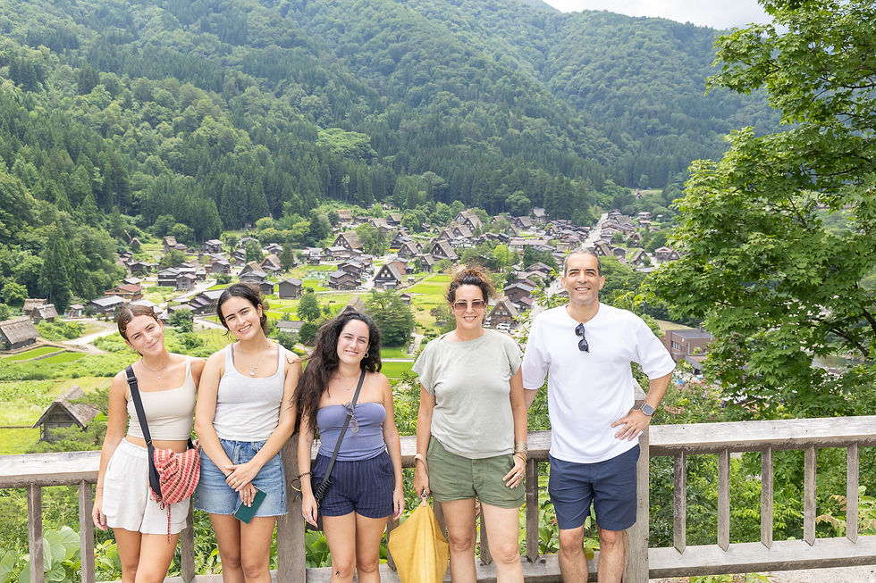 Five people smiling in front of a scenic village nestled in lush green hills, with traditional houses in the background. Bright, sunny day.