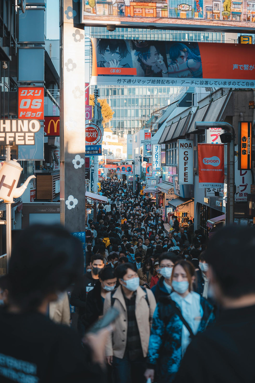 Crowd in masks fills a busy street lined with colorful signs, including cafes and stores, creating a vibrant urban scene in daylight.