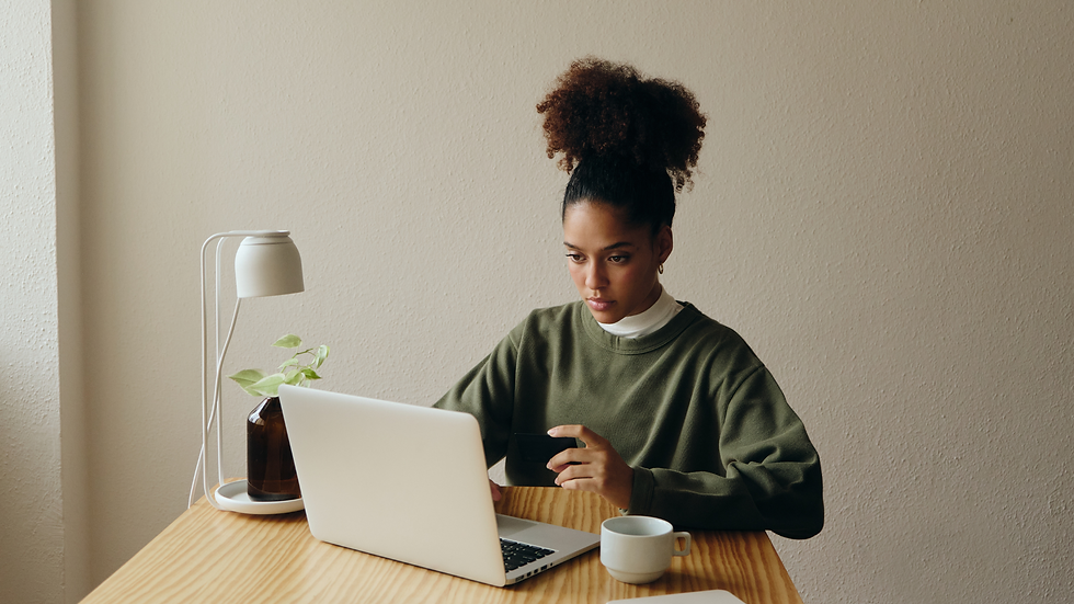 A woman in a green sweater uses a laptop at a wooden table, holding a card. A lamp and plant are in the background, creating a focused mood.