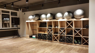 Moderner Kursraum mit Holzfußboden, Regalwand mit neatly sortierten Yoga-Matten, Hanteln und Gymnastikbällen. Das Design wirkt offen und minimalistisch, ideal für Yoga, Pilates oder funktionelles Training.
