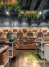 Präsentationswand im Sallys Welt Store mit Holzrückwand und dekorativen Regalen, die hochwertige Küchenaccessoires, Geschirr und Kochutensilien zeigen. Im Vordergrund stehen mobile Verkaufstische mit weiteren Produkten. Über der Szene hängen grüne Pflanzen von der Decke und verleihen dem modernen Interieur mit dunkler Decke und warmem Holzboden eine gemütliche, einladende Atmosphäre.