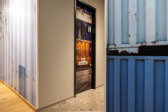 Flurbereich im Industrial Style mit rustikaler Containerverkleidung, kreativ gestalteter Tür mit gelbem Hafenmotiv und Schriftzug ‚HALT, wenn Tür in Bewegung‘ – markantes Designstatement im Büro