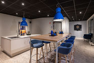 Moderner Küchen- und Loungebereich mit großer Holztafel, blauen Barhockern, auffälligen Industrieleuchten, weißer Kücheninsel und gemütlicher Sitzecke mit Dartboard – stilvoller Ort für Austausch und Pause