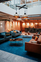 Moderner Loungebereich mit blauen und braunen Sofas, runden Couchtischen, Backsteinwand und großen Hängelampen im Industrial-Stil.