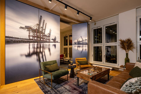 Moderne Büro-Lounge mit großem Hafenmotiv an der Wand, gemütlichen Designmöbeln in warmen Farben, großem Glastisch und dekorativen Details wie Trockenblumen und Spotlights – urban-maritime Wohlfühloase