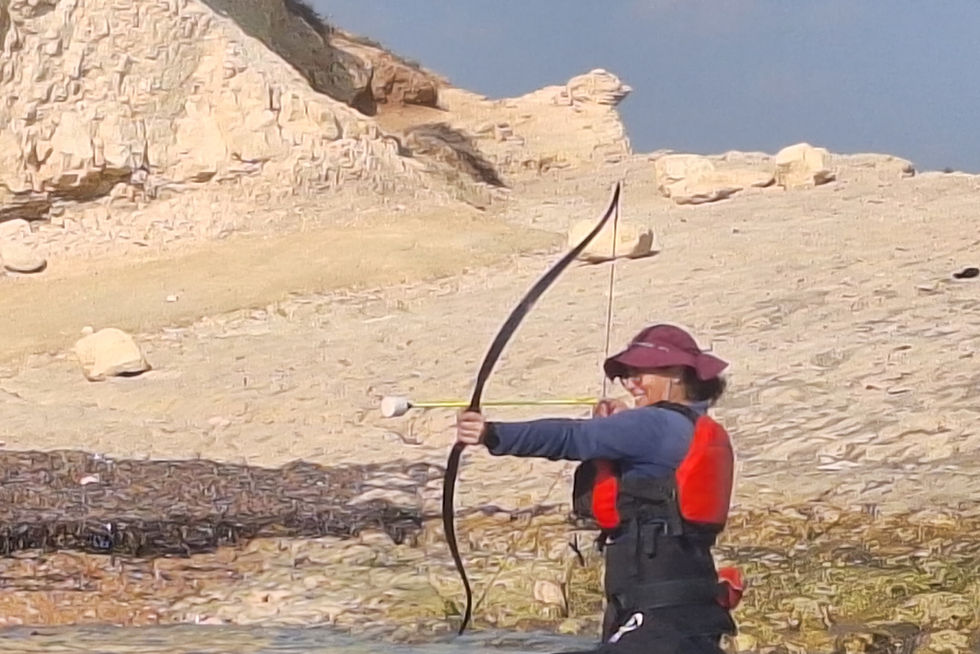 Lella, in red life vest and hat aims a bow on rocky shore. Clear sky and textured cliffs in the background. Appears focused.