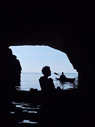Silhouetted sea kayakers viewed from inside a coastal cave near St Paul's Island, Malta, during a guided sea kayaking adventure. The dark cave interior frames the bright Mediterranean Sea beyond, where paddlers rest on calm water beneath the cave entrance. Reflections shimmer across the sheltered water while the natural limestone walls create a dramatic contrast between light and shadow. Accessible only from the sea, caves such as this provide sea kayakers with a unique perspective of Malta's coastline and geological formations. The image captures the tranquillity, exploration and sense of discovery experienced while paddling through hidden coastal features and remote sections of shoreline. Guided sea kayaking allows visitors to explore caves, coves and natural landmarks while enjoying Malta's crystal clear waters and spectacular coastal scenery from water level.