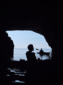 Silhouetted sea kayakers viewed from inside a coastal cave near St Paul's Island, Malta, during a guided sea kayaking adventure. The dark cave interior frames the bright Mediterranean Sea beyond, where paddlers rest on calm water beneath the cave entrance. Reflections shimmer across the sheltered water while the natural limestone walls create a dramatic contrast between light and shadow. Accessible only from the sea, caves such as this provide sea kayakers with a unique perspective of Malta's coastline and geological formations. The image captures the tranquillity, exploration and sense of discovery experienced while paddling through hidden coastal features and remote sections of shoreline. Guided sea kayaking allows visitors to explore caves, coves and natural landmarks while enjoying Malta's crystal clear waters and spectacular coastal scenery from water level.