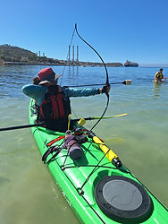 Lella practicing kayak archery during a guided sunset adventure in Malta, aiming a bow while seated in a green sea kayak on calm crystal-clear Mediterranean waters. Surrounded by scenic coastline and open sea views, this unique outdoor activity combines sea kayaking and archery in a safe and exciting environment led by certified instructors from GOAK and AIM. The experience highlights balance, focus, coordination, and adventure tourism while enjoying Malta’s beautiful coastal scenery. Perfect for beginners and experienced paddlers looking for a memorable sunset activity on the water with kayaking, archery, and outdoor exploration.