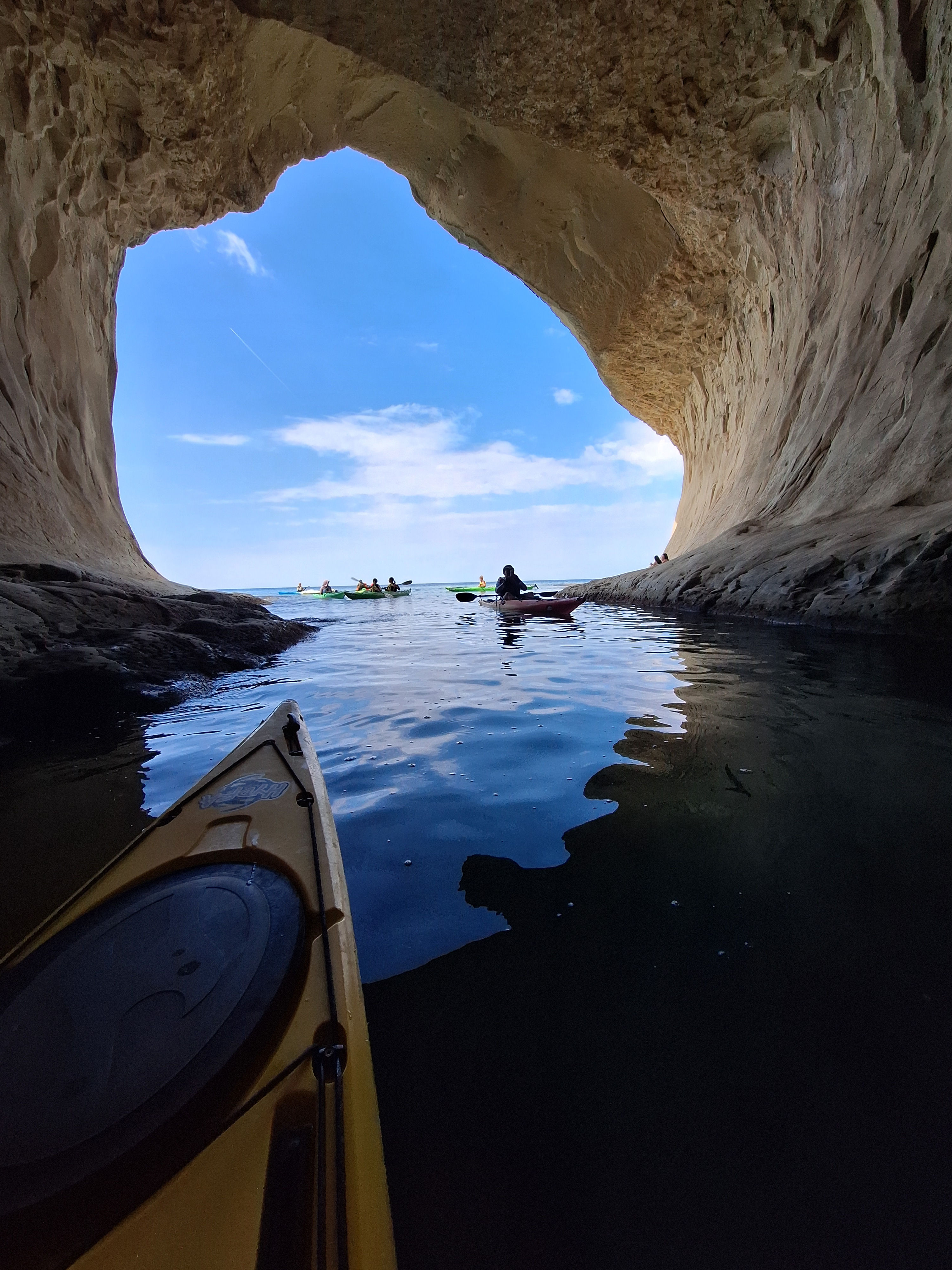 View from inside Ta' Marija Cave near Ä nejna, Malta, looking out through a spectacular natural limestone opening towards the bright blue Mediterranean Sea. The bow of a sit-inside sea kayak is visible in the foreground, with other kayakers paddling outside the cave during a guided adventure with Get Out and Kayak Malta.
