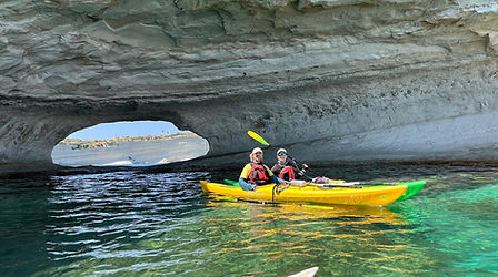 Two paddlers in a yellow tandem sea kayak explore the crystal clear waters of Il-Ħofra, Malta, during a guided sea kayaking adventure. The kayak passes beneath a dramatic limestone overhang featuring a natural opening carved by centuries of wave action along Malta's rugged southeastern coastline. Sunlight illuminates the turquoise Mediterranean water, revealing the striking geology and natural beauty that make Il-Ħofra a favourite destination for sea kayakers. Accessible primarily from the sea, this hidden coastal feature offers paddlers a unique perspective of Malta's cliffs, rock formations and marine environment. The image captures the adventure, exploration and tranquillity of discovering remote coastal landscapes by kayak while enjoying calm conditions and spectacular scenery with certified and registered Paddle UK Sea Kayak Guides.