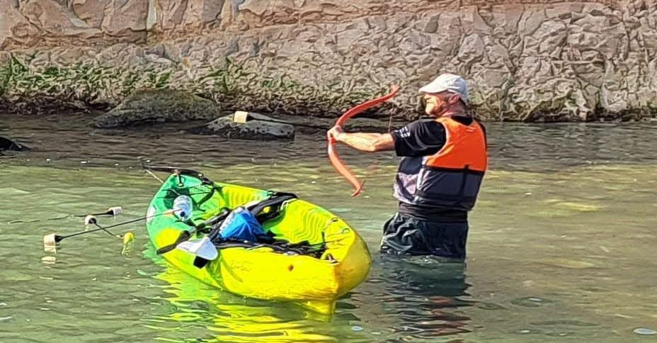 Man in water beside a neon green kayak, wearing an orange life jacket, holds a bow and arrow for kayak archery. Rocky wall in background, clear day.