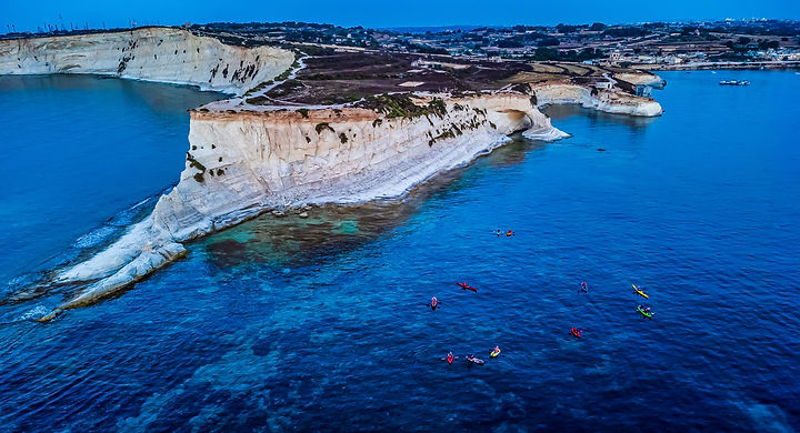 Drone photograph showing sea kayakers exploring the limestone cliffs and turquoise waters of the Munxar coastline in Malta.
