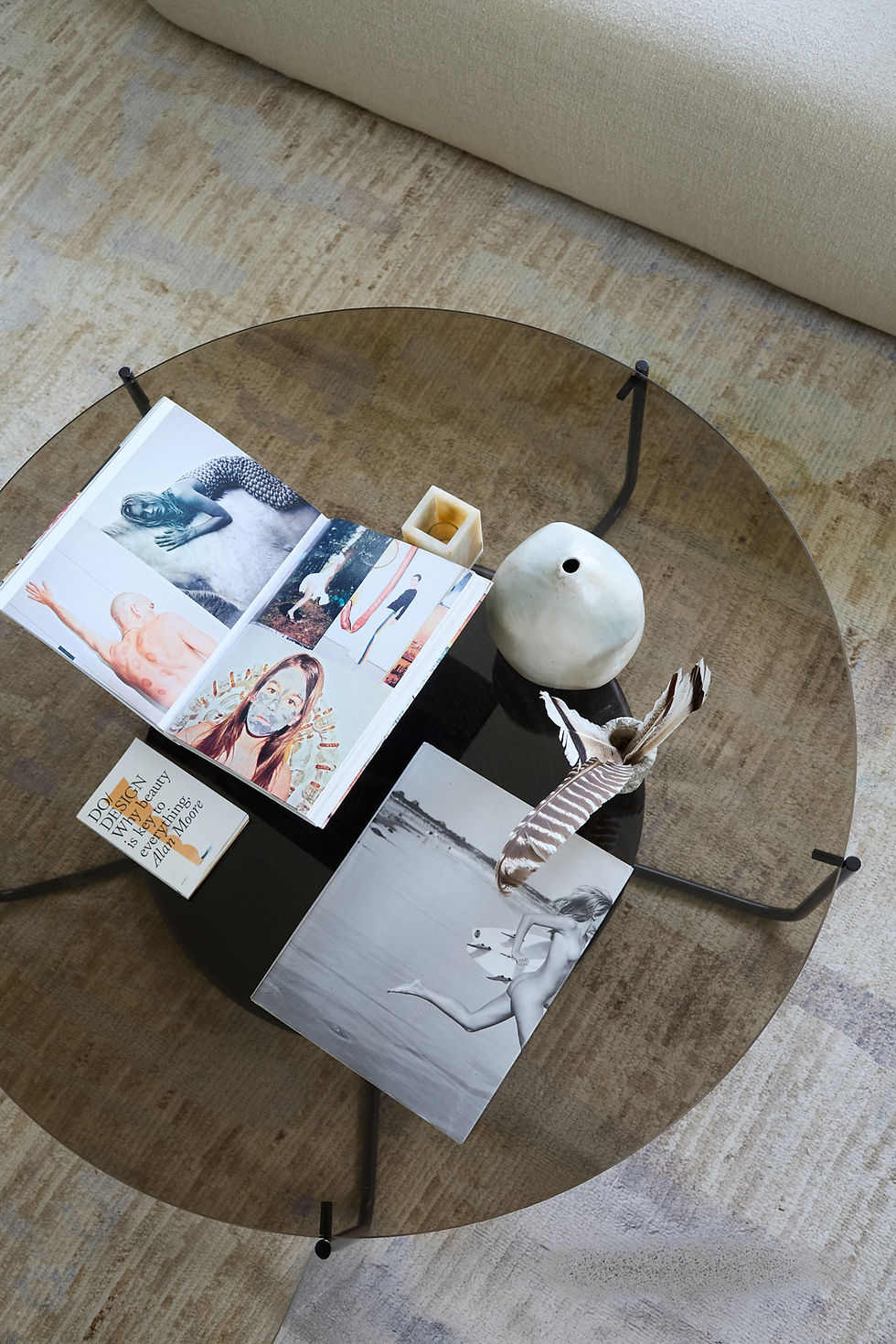 Glass coffee table with art book, feather, candle, and ceramic vase on beige rug. Book titled "Do Design: Why Beauty..." is visible.