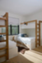 Bunk beds in a bright room with wooden ladders and blue-gray pillows. A window with a view of trees and a small stool with plants add charm.