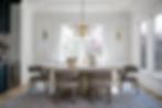 Elegant dining room with round table, six gray chairs, modern chandelier, and large windows. White walls, soft lighting, and calm ambiance.