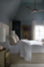 Cozy bedroom with soft white bedding, two cream pillows, a dark ceiling fan, and framed art on muted blue walls. Sunlight filters through linen draperies.