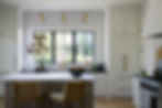 Bright kitchen with white cabinets and a large window. A bowl of fruit and plants sit on the island. Golden fixtures, cozy and inviting.