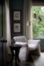 Cozy room with a fluffy chair, black side table with photos, floor lamp, abstract art on walls, green view outside, Belgian linen draperies.