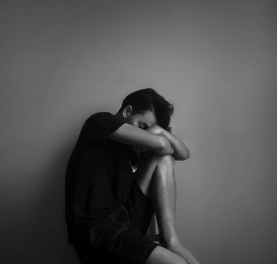 A black and white image of a person seated on a chair against a plain wall. Wearing a dark t-shirt and shorts, their head rests on their arms, crossed on one knee. The somber atmosphere hints at depression, with the person appearing deep in thought, withdrawn, and sad.