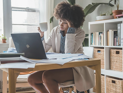 A person with curly hair sits at a cluttered wooden table, holding a white mug and looking at a laptop screen during a therapy session for anxiety. Papers are scattered on the table and floor, while shelves with books and plants provide a calming backdrop. The person wears a light blazer, striped shirt, and rust-colored socks.