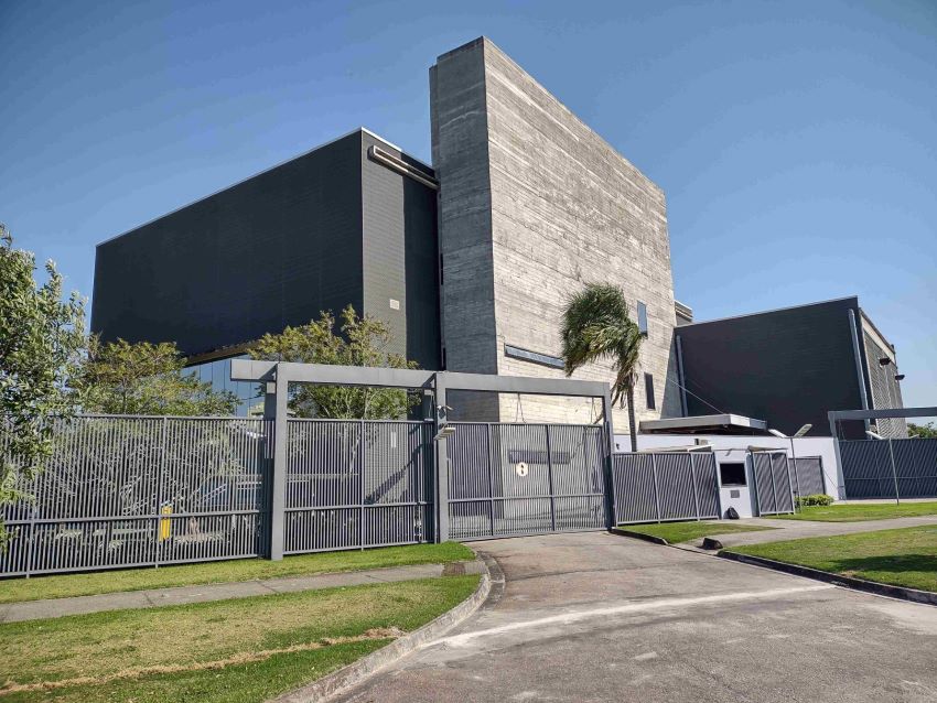 Prédio em concreto visto de longe, atrás de portão e grades