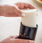 Eine Frau mit elegant manikürten Fingern und funkelndem Ring setzt eine cremefarbene Stumpenkerze in ein schwarzes Glasgefäß mit der Aufschrift Barowski candle couture ein. Die Szene vermittelt den Eindruck von Qualität und Sorgfalt bei der Handhabung der hochwertigen Barowski Kerzen, die durch ihr edles Design und luxuriöses Erscheinungsbild überzeugen.