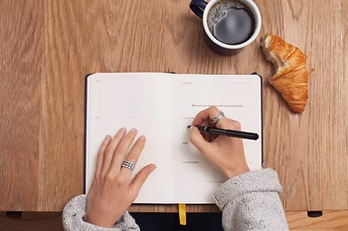 Hands writing in an open planner on a wooden table, with a coffee cup and croissant nearby; calm morning workspace.