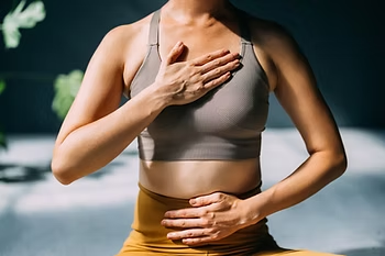 Woman in gray sports bra sits meditating, one hand on chest and one on abdomen, in a calm indoor yoga setting.