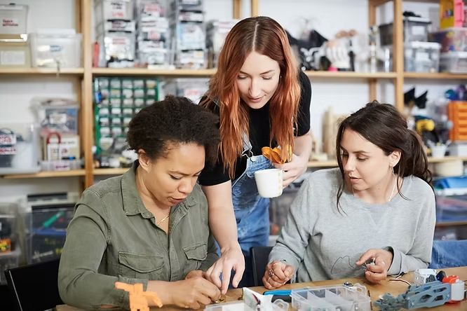 Three women collaboratively work on a project at a table; one holds a mug. Shelves packed with supplies fill the background.