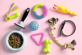 Assorted colorful dog toys and a bowl of kibble on a pink background, arranged in a neat flat lay.