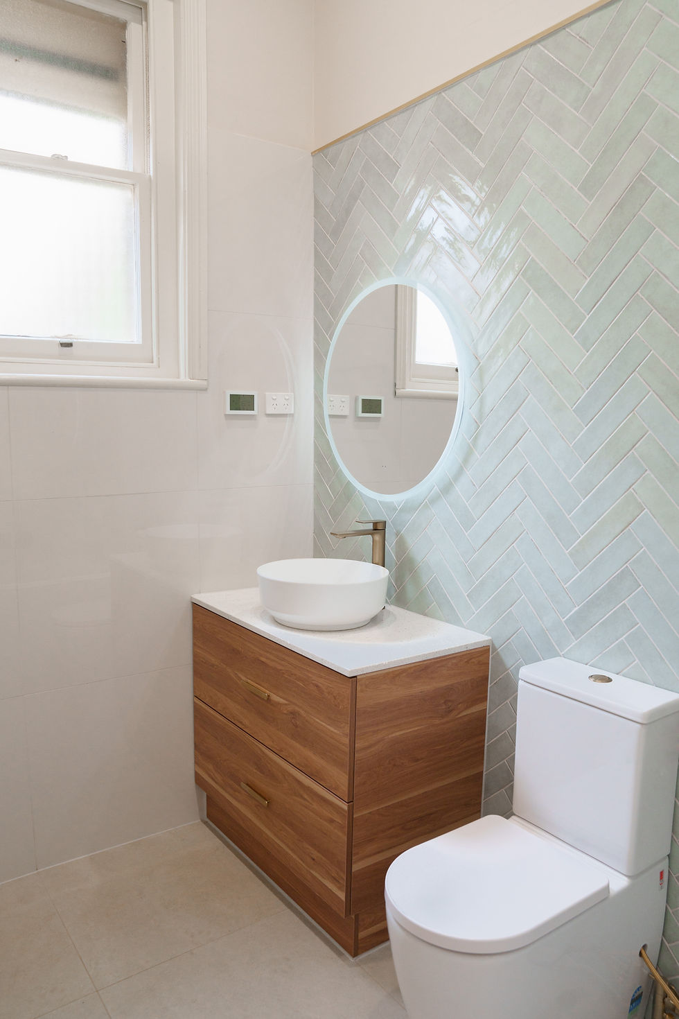 A modern bathroom features a sleek wooden vanity with a minimalist basin, complemented by herringbone patterned tiles and a round LED mirror, combining style and functionality.