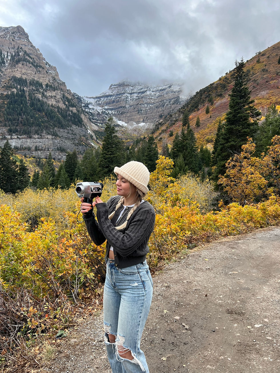 A person in a knit hat films with a Super 8 Video camera against a backdrop of mountains and autumn trees. The mood is serene and adventurous. It is in Provo Utah, Alpine Loop