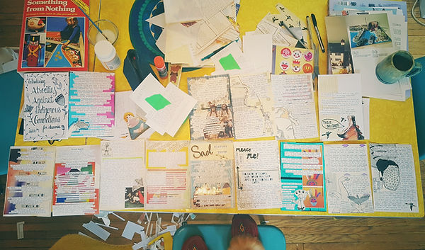 This is an overhead shot of my incredibly messy desk, halfway through the production phase of the first issue of "Atrocities Against Indigenous Canadians for Dummies." There are papers and zine pages everywhere as well as my coffee cup, my moccasins in the whot. My table is a yellow, 1950's table and my chairs are chrome with blue vinyl