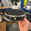 Thumbnail: Side and top view of a practice tamborim with boxes of percussion instruments in the background.