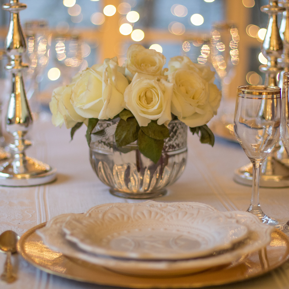 Elegant table setting with white roses in a glass vase, surrounded by silver candlesticks and glassware. Warm, festive bokeh lights glow.