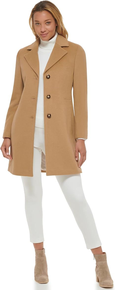 Woman in a tan coat over a white turtleneck and pants, standing confidently. She wears tan boots. White background.