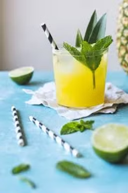 Yellow citrus drink in a glass with mint and a striped straw, on a blue table with lime halves and striped straws.
