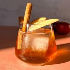 A glass of amber-colored drink with ice, apple slices, and a cinnamon stick. Warm lighting on a peach surface; apples in the background.