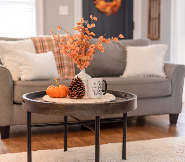 Cozy living room with a table holding a pumpkin, pinecone, a mug with text, and orange leaves. Gray couch with plaid blanket behind.