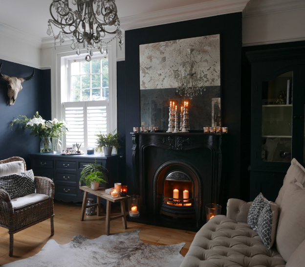 Cozy living room with a dark fireplace, lit candles, chandelier, and plants. Elegant decor, natural light from a window, serene mood.