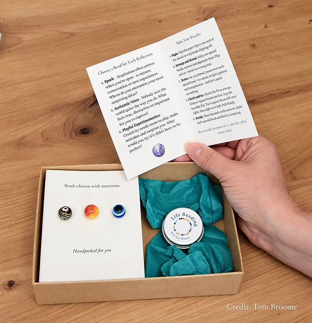 An opened Creatitivy! kit with orange, stripey and blue beads, a Life Beading tin and a hand holding open the guide card with three prompts and instructions for bracelet making.