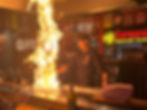 A captivating action shot of the chef performing, mid flip, the fiery grill illuminating their face, embodying the energy and excitement of Teppanyaki cooking