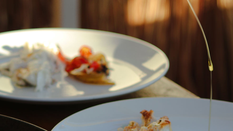 A hand pours oil on grilled fish and veggies on a white plate, set on a wooden table with an earthy-toned background, creating a rustic feel.
