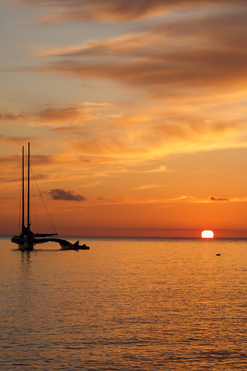 A catamaran floats on calm water at sunset. The sky glows with orange and pink hues, creating a serene and tranquil scene.
