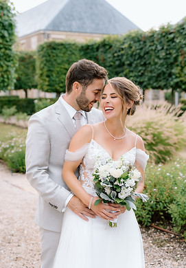 Ein lachendes Brautpaar im Garten des Château de Varennes: Der Bräutigam umarmt die Braut liebevoll von hinten, während sie einen weissen Blumenstrauss hält. Im Hintergrund sind gepflegte Grünflächen und die eleganten Gebäude des Châteaus zu sehen
