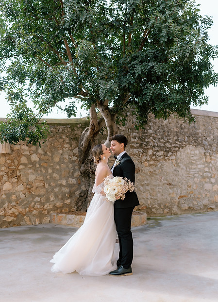 Braut und Bräutigam umarmen sich vor einer rustikalen Steinmauer und einem grossen Baum in der Finca Morneta auf Mallorca. Die Braut trägt ein leichtes, fliessendes Kleid und hält einen cremefarbenen Blumenstrauss, während beide einander liebevoll anlächeln