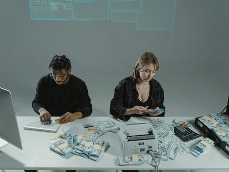 Overhead view of two people at a table surrounded by stacks of cash, a bill counter, and credit cards in front of a computer monitor, representing the financial risk and fraud enabled by AI customer service chatbot vulnerabilities