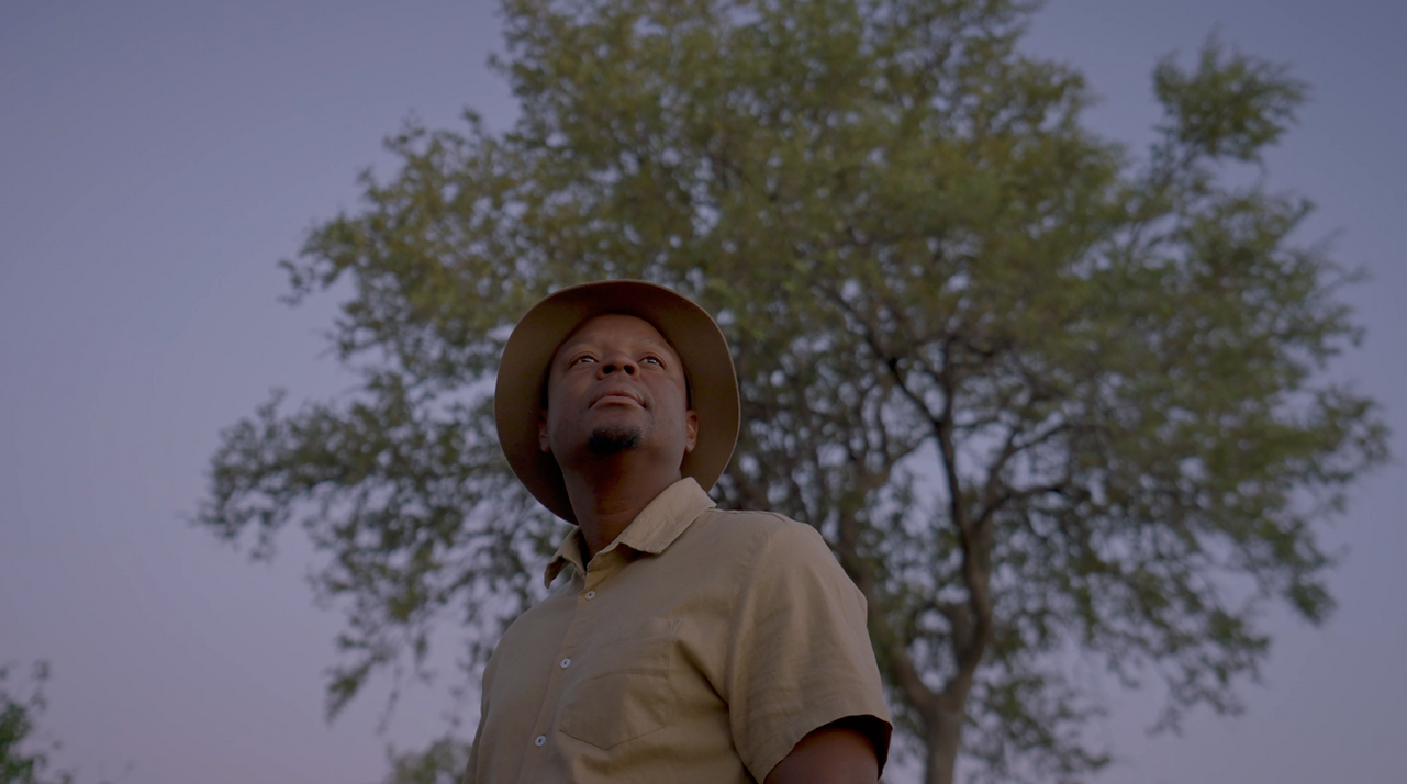 Color-graded shot of a man wearing a safari hat, standing beneath a tree in an African landscape.